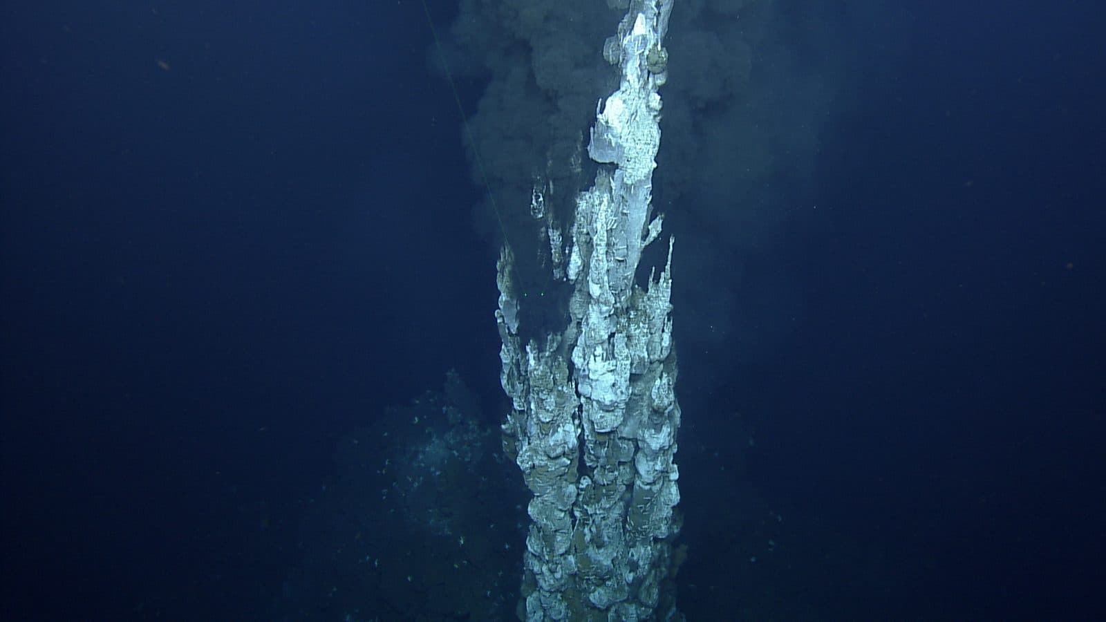 A tall pale hydrothermal chimney rising from the seafloor into dark blue water, with black smoke pouring from its upper part