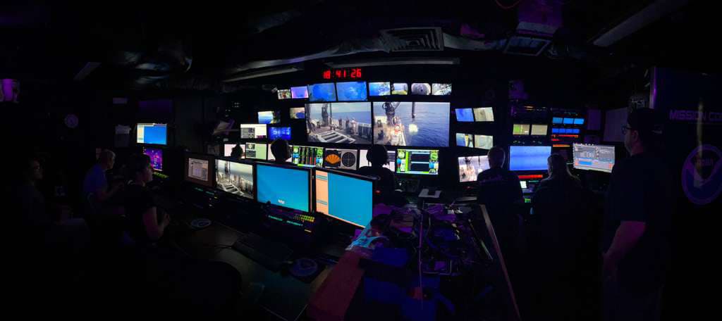A darkened shipboard control room, a wall of monitors showing live seafloor video, sonar and navigation data, with the team watching from two rows of consoles