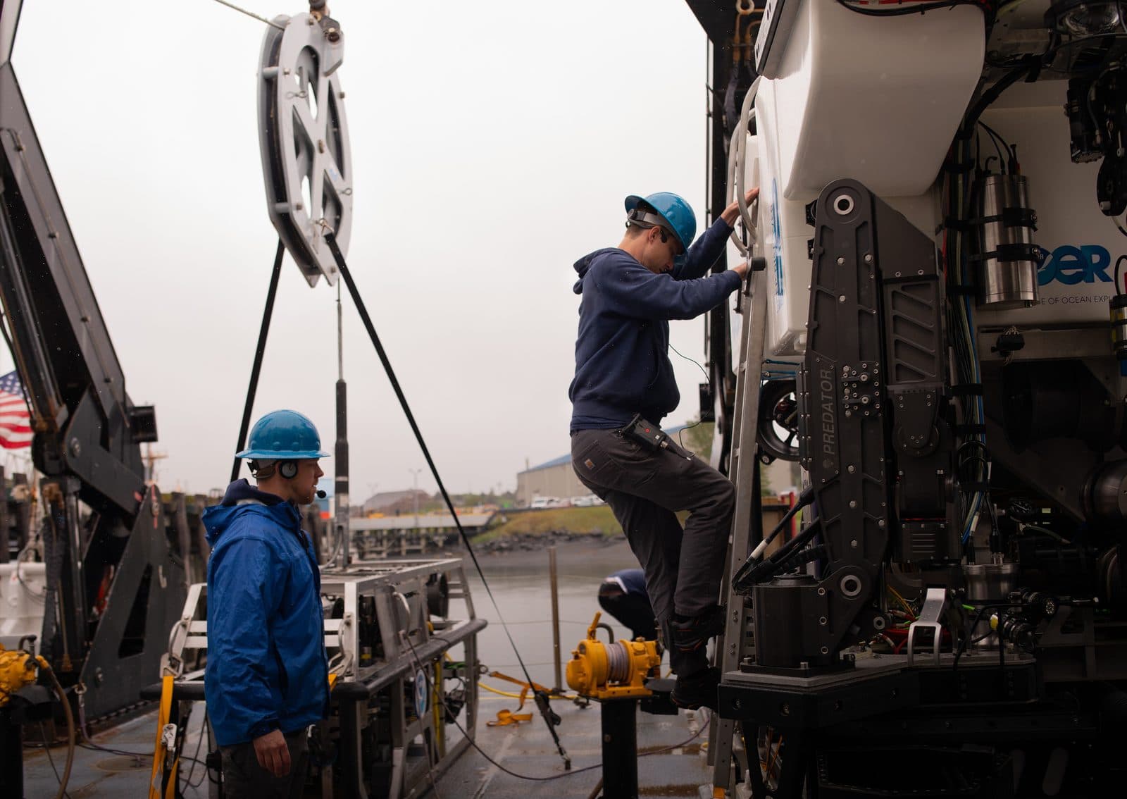 Two engineers in blue hard hats working on a large white remotely operated vehicle on a ship's deck